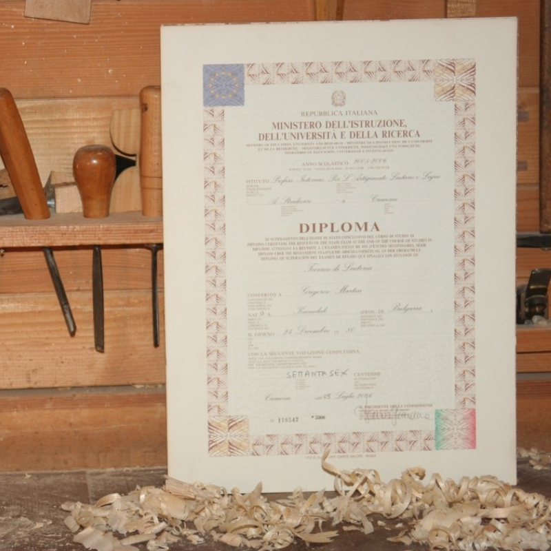 Framed diploma leaning against a wooden workshop wall with wood shavings on the floor and chisels nearby.