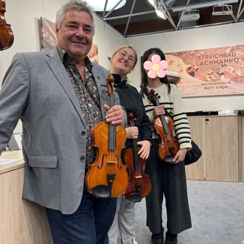 Three people pose with violins in a showroom, smiling at the camera, with a display and banner in the background.