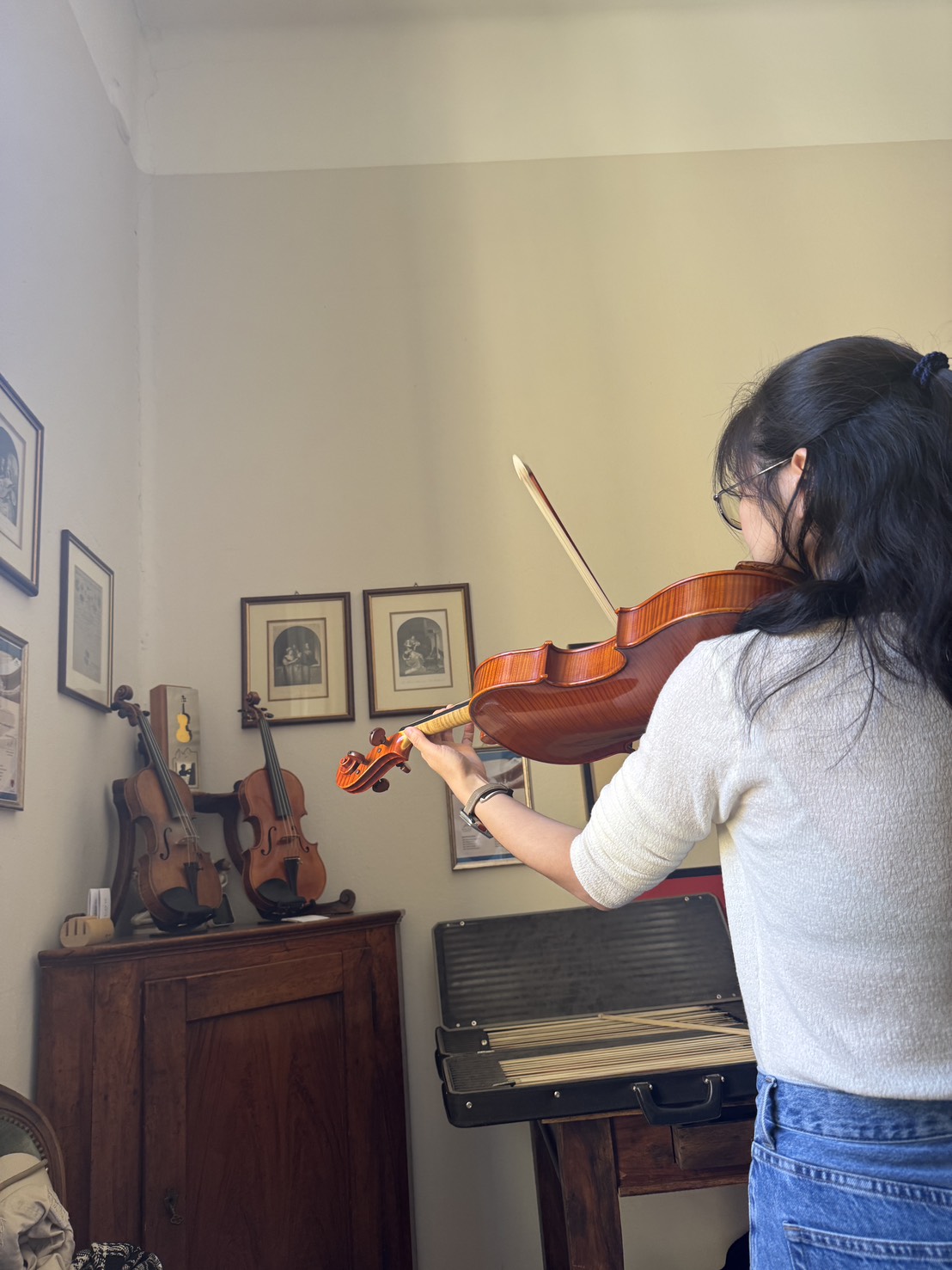 S226263080 | 希爾提琴 HEL Violins Person playing a wooden violin in a cozy room, standing at a music stand with a violin case nearby.