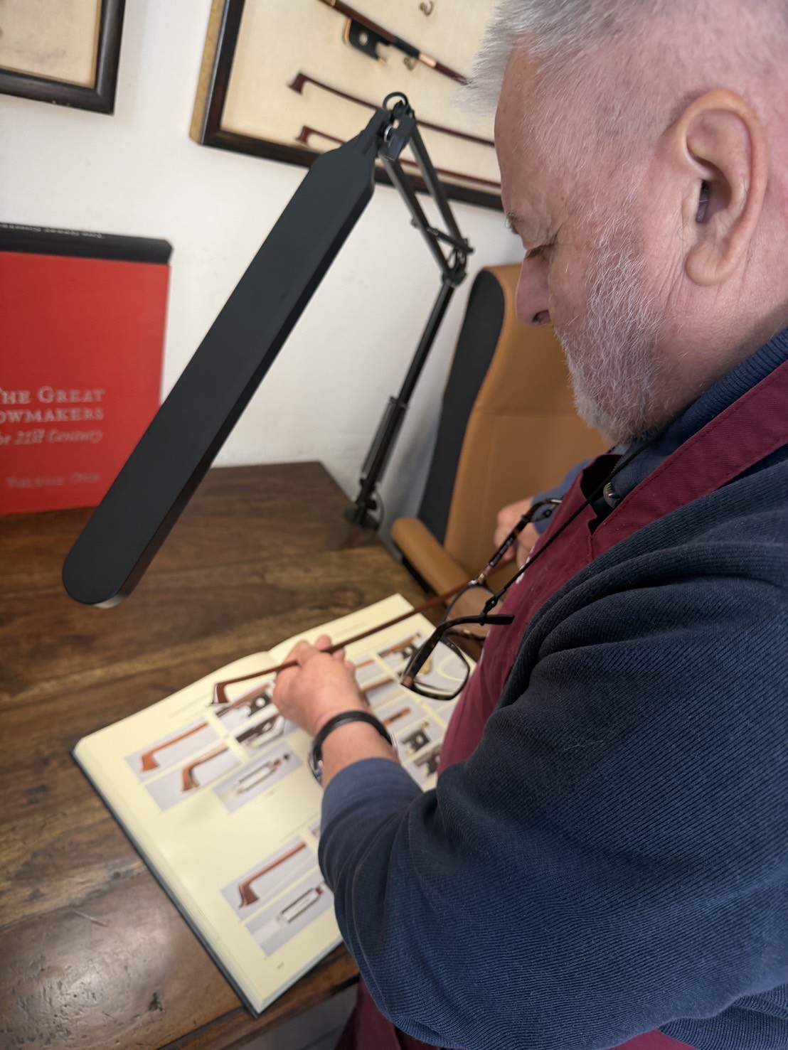 S22626309 | 希爾提琴 HEL Violins Older man with gray beard and glasses reviews a photo album at a wooden desk, with a large adjustable lamp arm in the foreground.